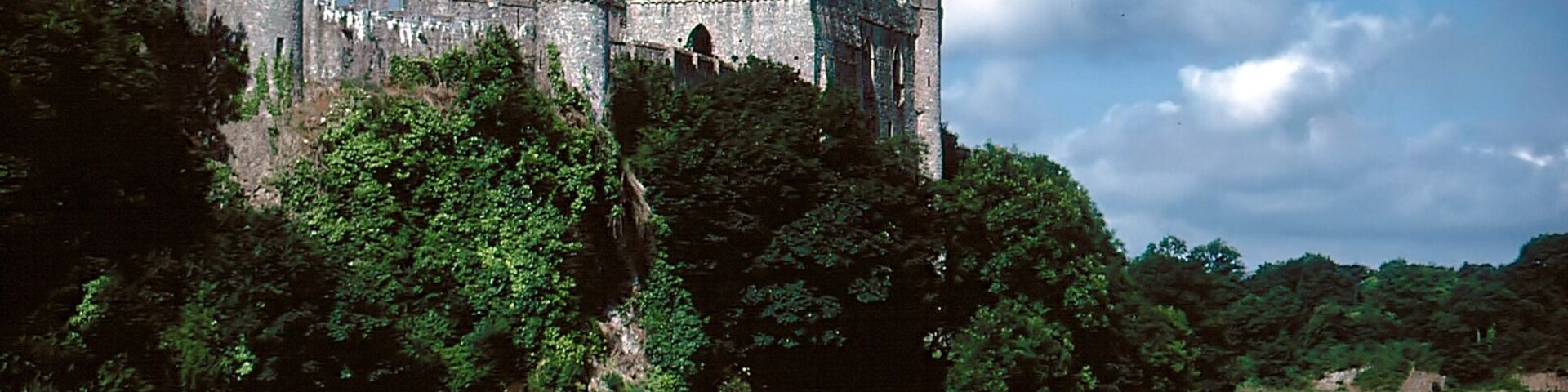 Um castelo histórico numa colina com vista para um lago calmo, rodeado por vegetação exuberante.