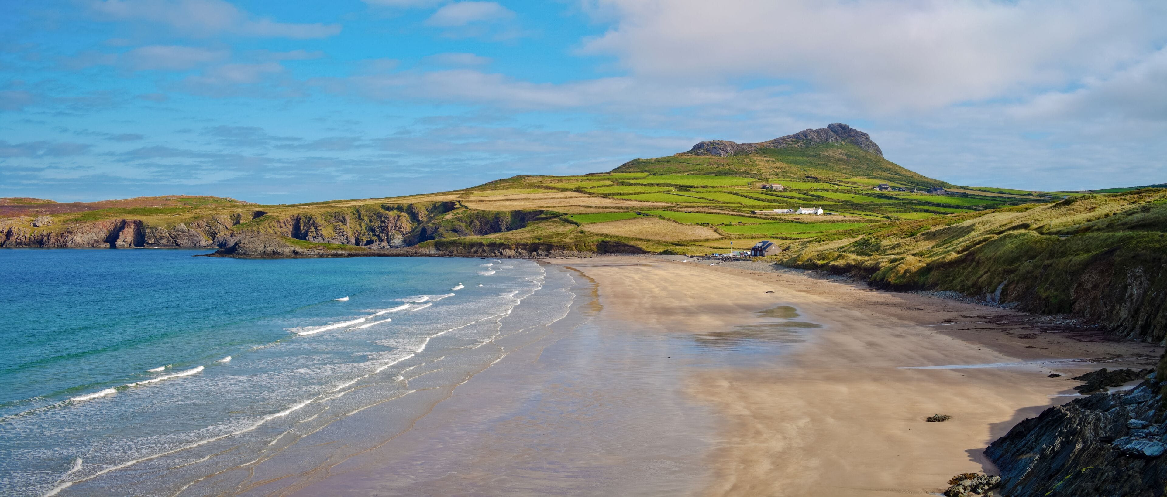 Un paisaje costero con una playa arenosa, colinas verdes y un acantilado rocoso.