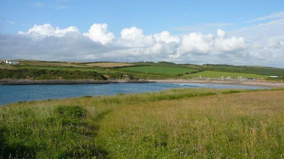 A coastal landscape with a grassy foreground, a body of water, and rolling hills in the distance.