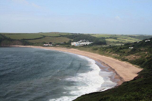 A coastal landscape with a sandy beach, rolling hills, and a white building in the distance.