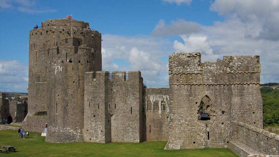 A historic stone castle with a prominent tower and battlements, surrounded by a well-maintained lawn and picnic tables.