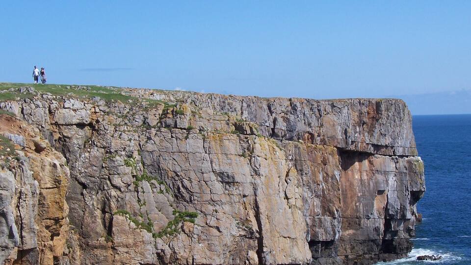 A rocky cliff with a small grassy area at the top where two people are standing.