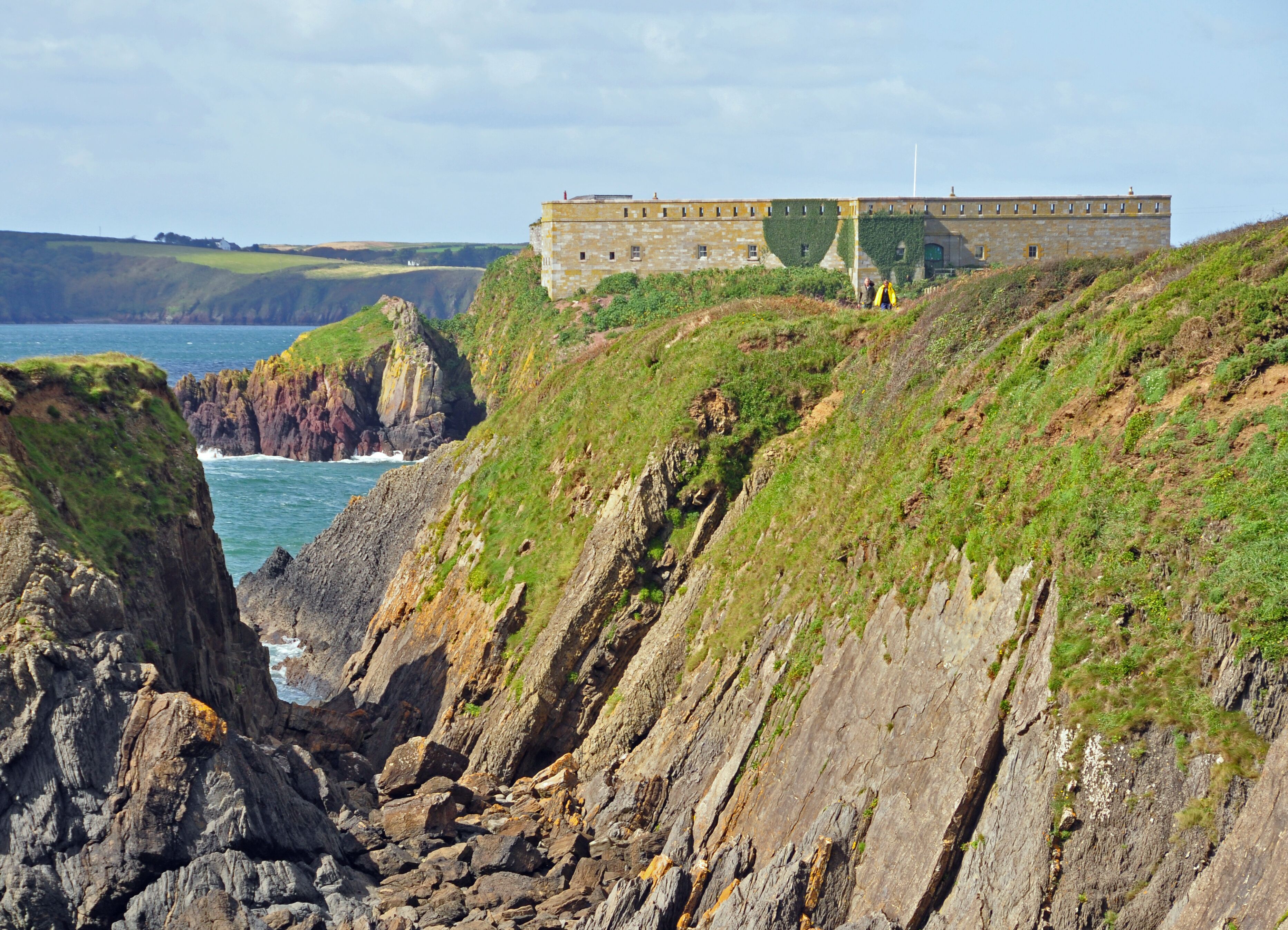 A coastal landscape with rocky cliffs, a building with ivy, and the ocean in the background.