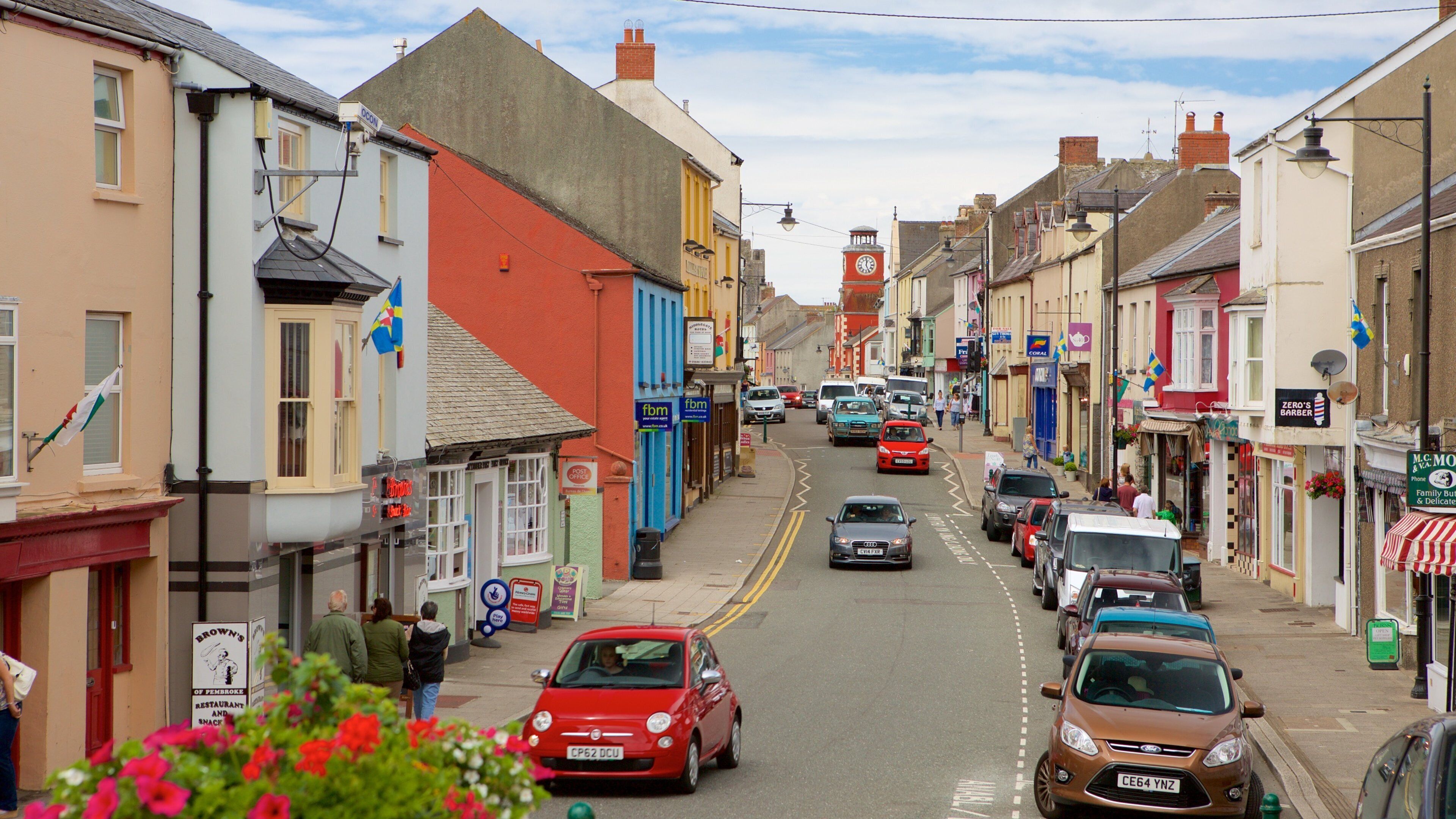 A street lined with colorful buildings, parked cars, and a clock tower in the distance.