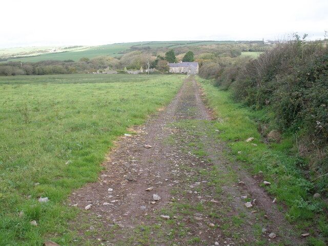 A rural dirt path leading to a stone house surrounded by fields and vegetation.
