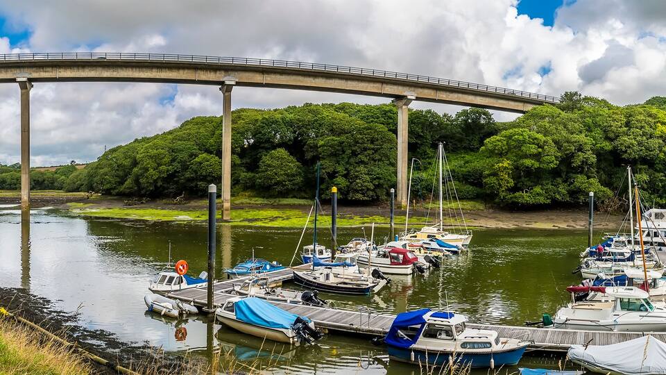 A river with boats docked at a pier, a bridge in the background, and a path alongside the river.