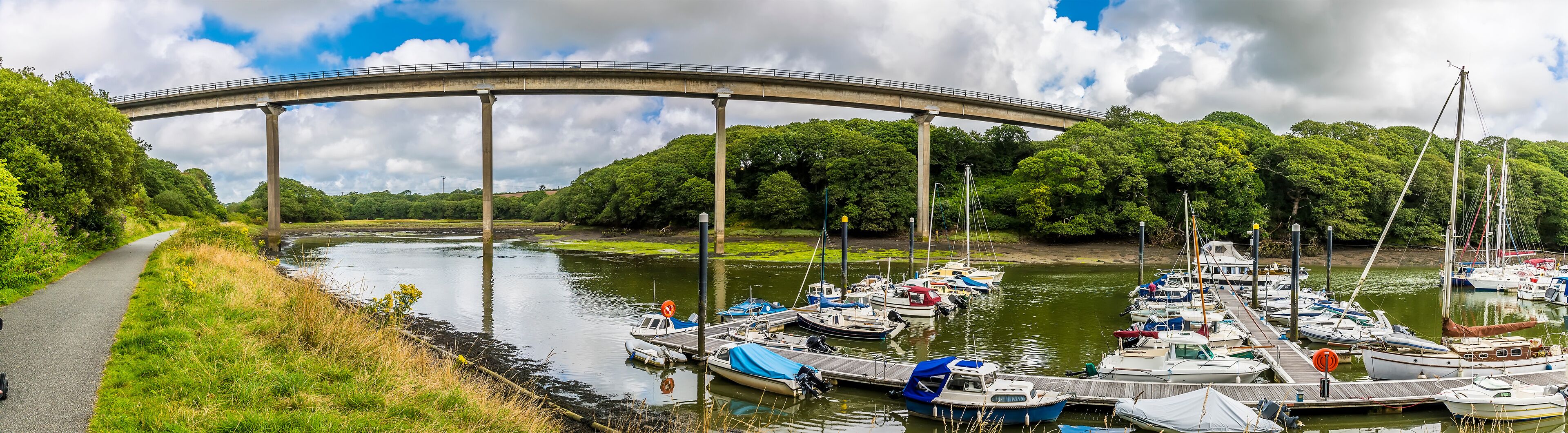A river with boats docked at a pier, a bridge in the background, and a path alongside the river.