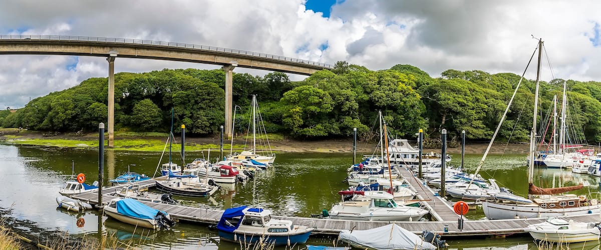 A river with boats docked at a pier, a bridge in the background, and a path alongside the river.