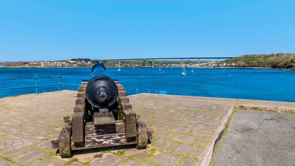 A cannon on a stone platform by a body of water with sailboats and a bridge in the background.