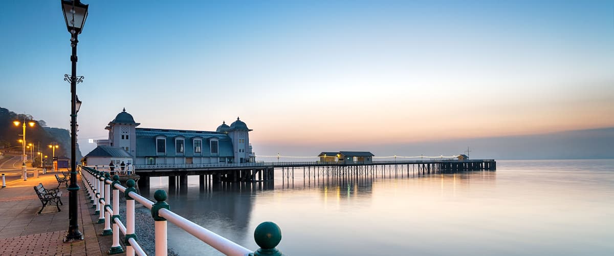 A calm waterfront with a pier extending into the water, a row of lamp posts, and a building with a dome and tower.