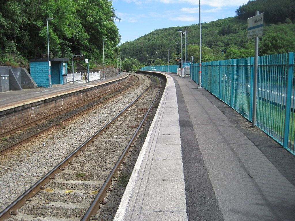 A railway station platform with tracks, a fence, and a hill in the background.