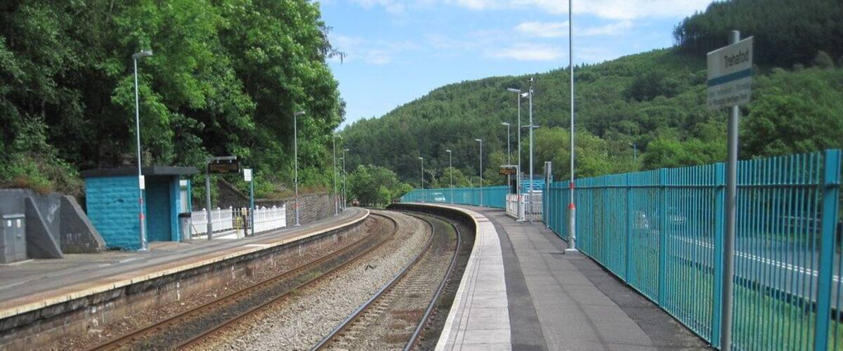 A railway station platform with tracks, a fence, and a hill in the background.