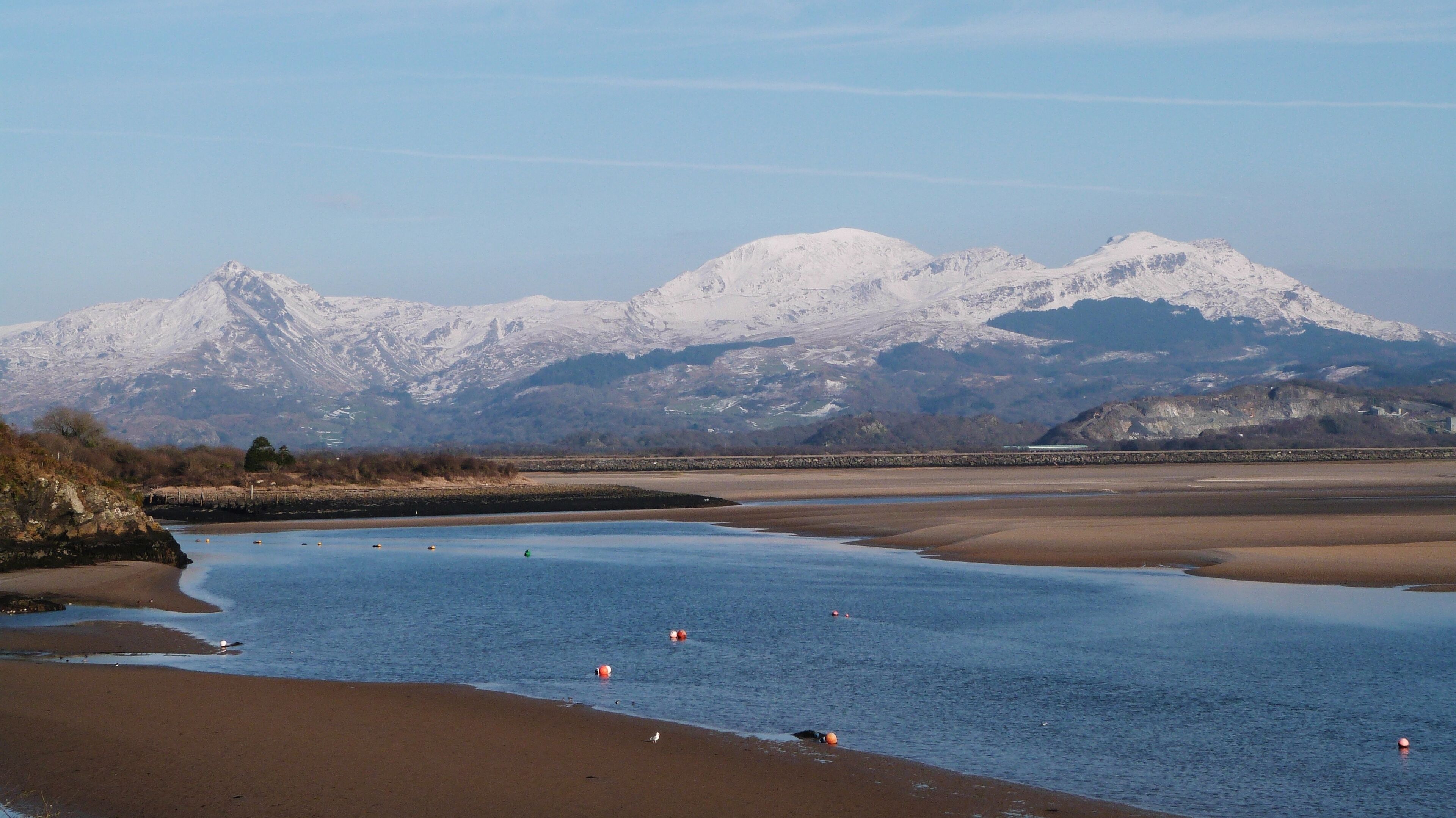 A coastal landscape with a sandy beach, calm water, and snow-capped mountains in the background.