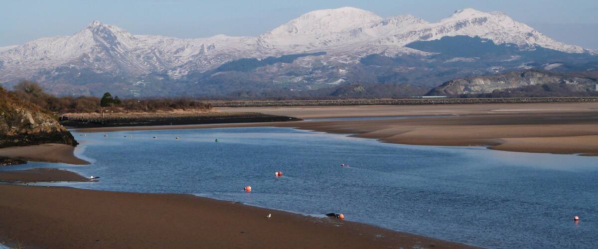 A coastal landscape with a sandy beach, calm water, and snow-capped mountains in the background.