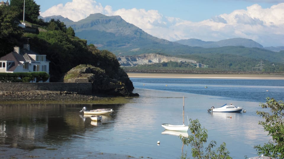 A coastal scene with calm water, boats, a house, and mountains in the background.