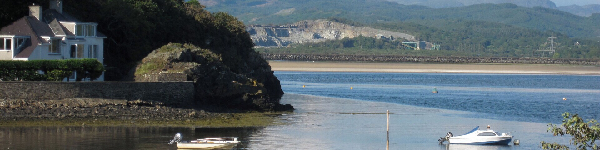 Escena costera con agua tranquila, barcos, una casa y montañas al fondo.
