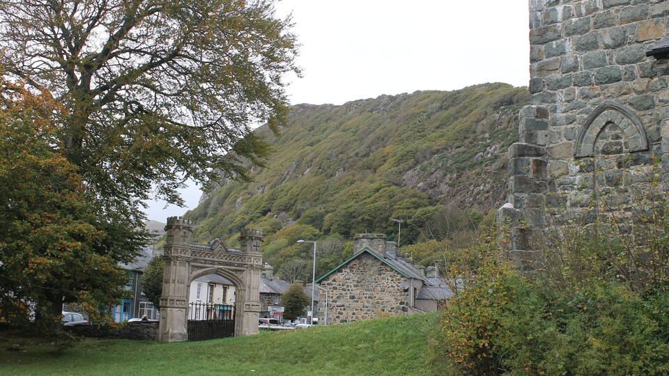 A stone church with a green roof, surrounded by lush greenery and a hill in the background.