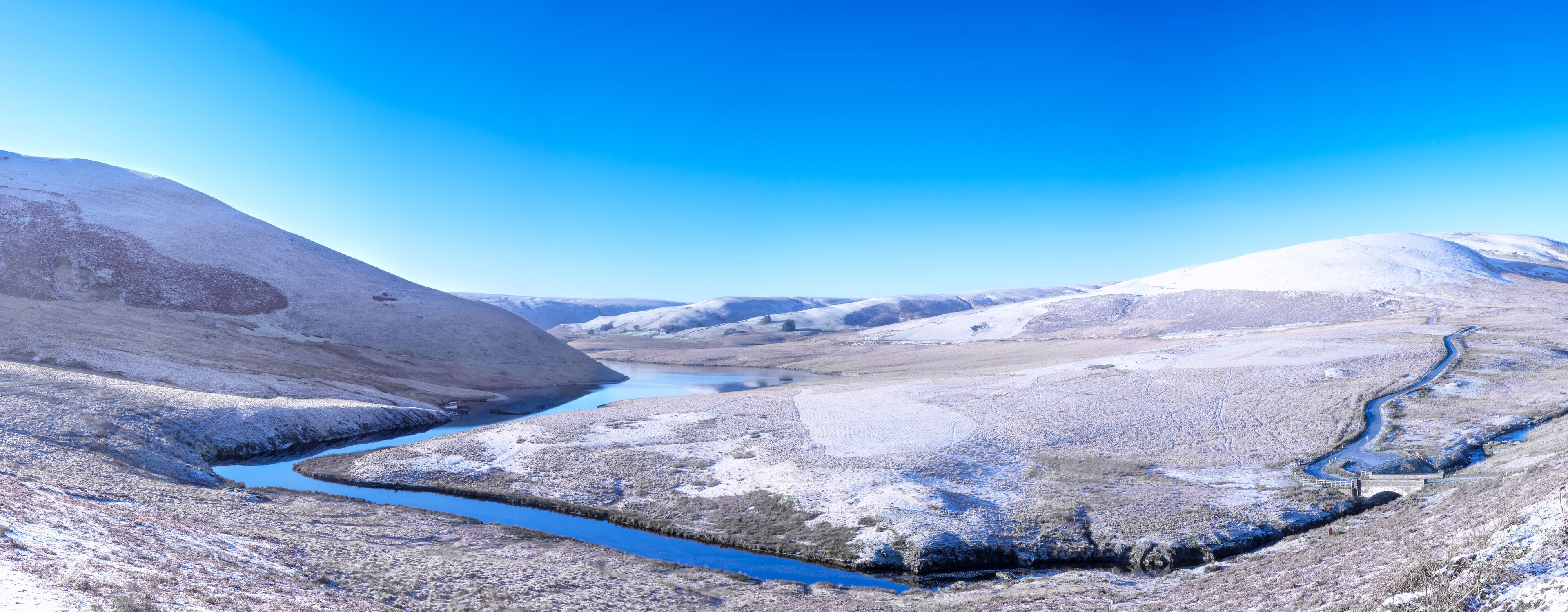 Elan Valley in Wales showing Afon Elan river flowing through a snowy panorama of a winter scene of mountains and blue sky.