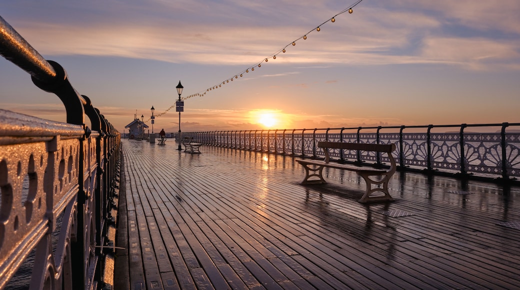 A wooden pier with benches and string lights, leading to a sunset over the water.