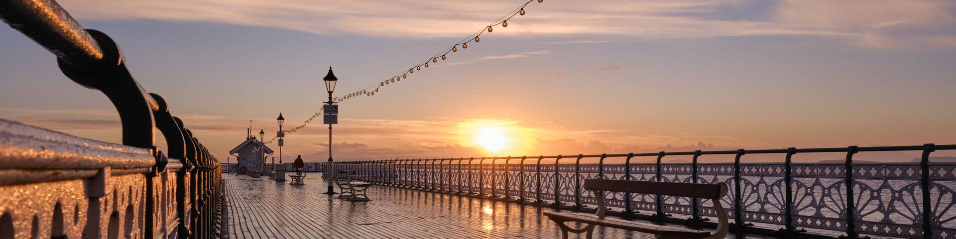 A wooden pier with benches and string lights, leading to a sunset over the water.