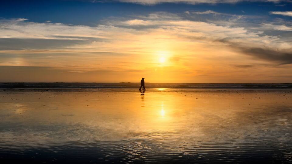A beach at sunset with a person standing in the center, reflecting the sun's rays on the wet sand.