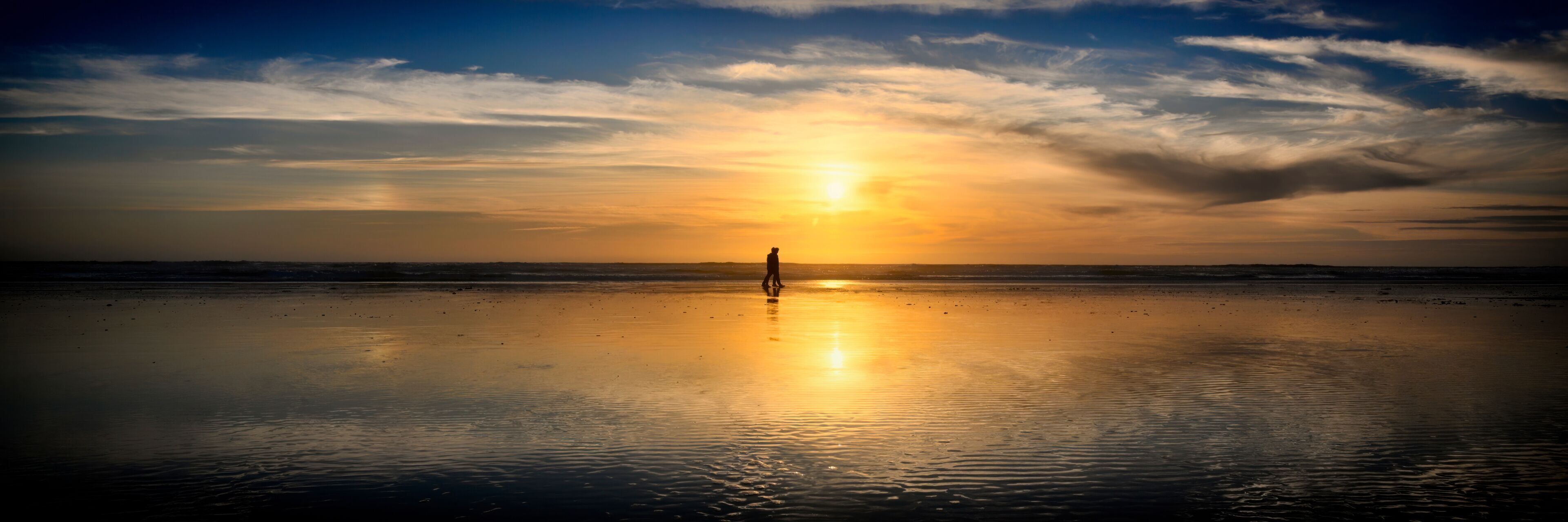 A beach at sunset with a person standing in the center, reflecting the sun's rays on the wet sand.