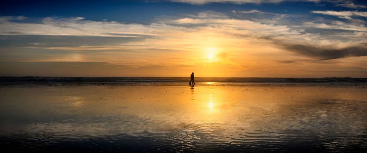 A beach at sunset with a person standing in the center, reflecting the sun's rays on the wet sand.