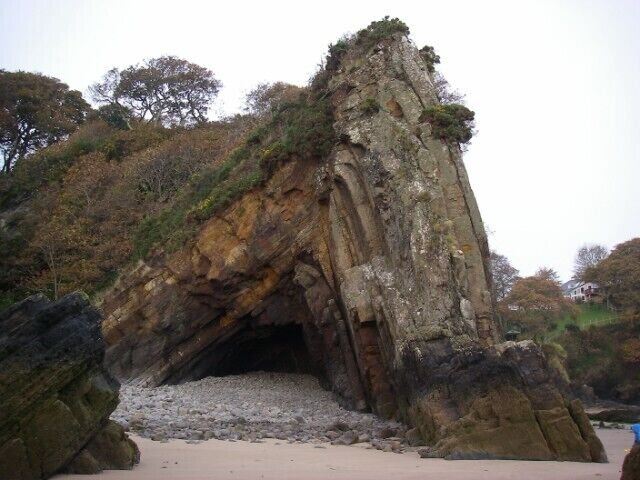 Uma grande formação rochosa com a entrada de uma caverna, rodeada por árvores e uma praia de areia.