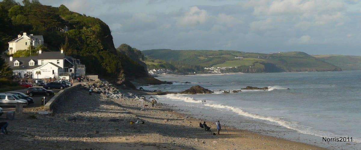 A coastal town with a rocky beach, a white building with a sign, and hills in the background.