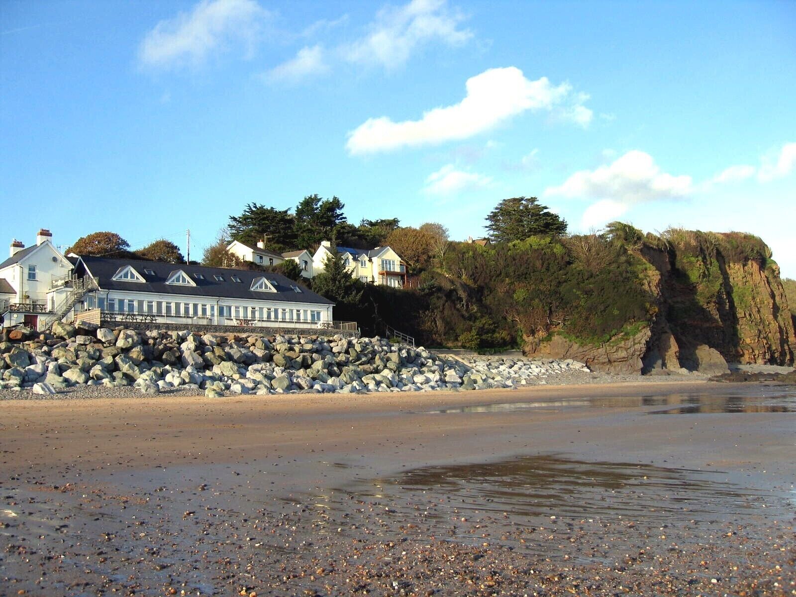 A coastal area with a large white building with a dark roof, a rocky shoreline, and a cliff with vegetation.