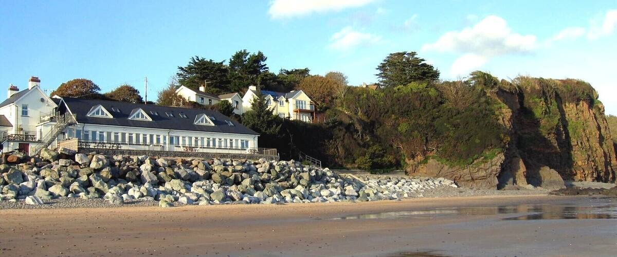 A coastal area with a large white building with a dark roof, a rocky shoreline, and a cliff with vegetation.