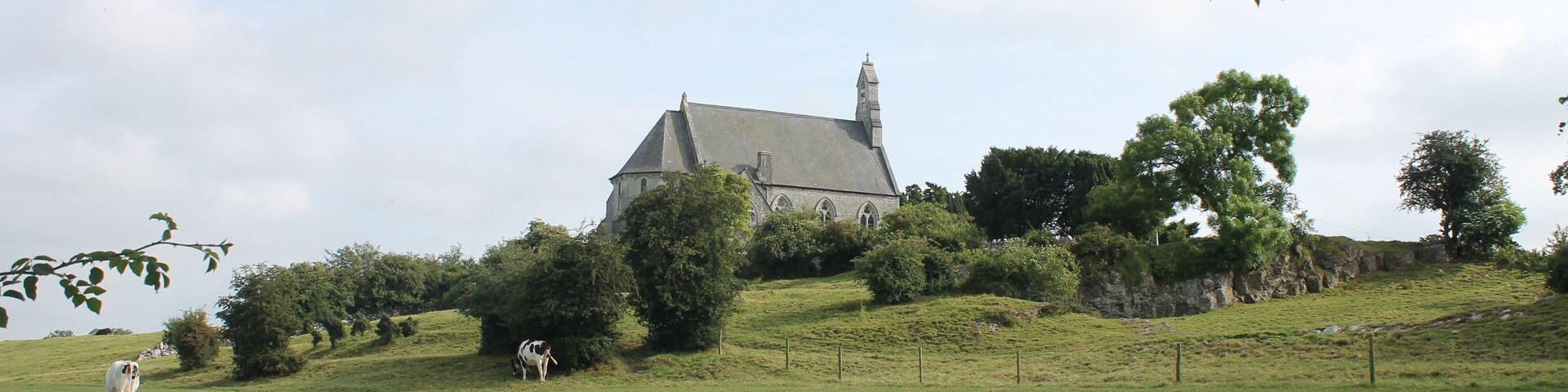 Paisagem rural com uma casa grande numa colina, rodeada por árvores e um campo relvado.