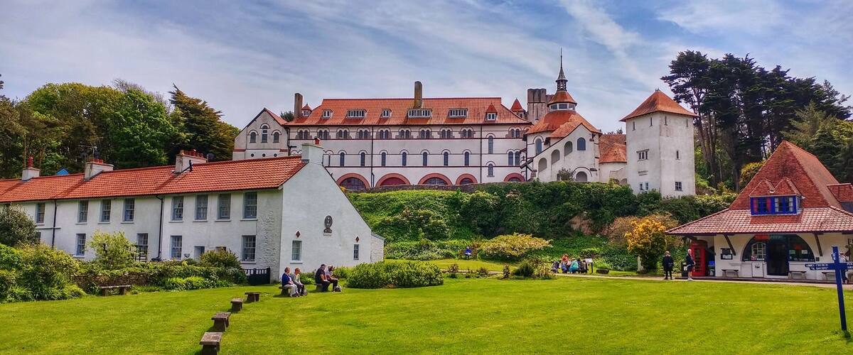 A castle with red-tiled roofs, white walls, and arched windows. There are benches in a grassy area in front of the castle.