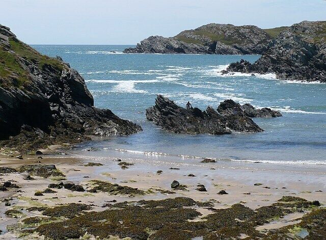 A rocky coastline with waves crashing onto the shore, a sandy beach, and seaweed-covered rocks.