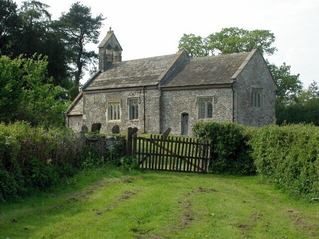 A stone church with a bell tower, surrounded by greenery and a wooden gate.