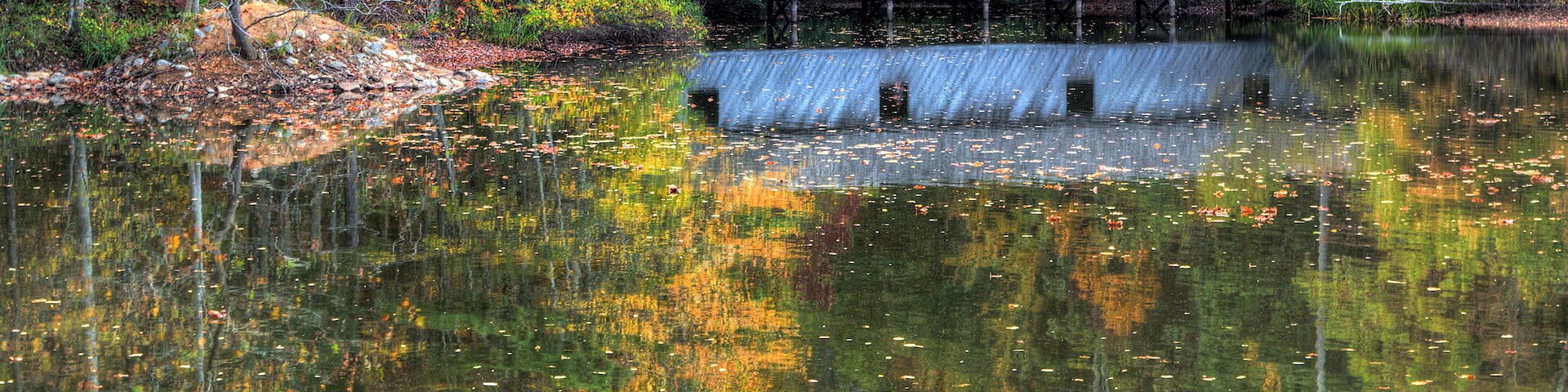 A rustic cabin on stilts by a calm lake surrounded by autumn-colored trees.