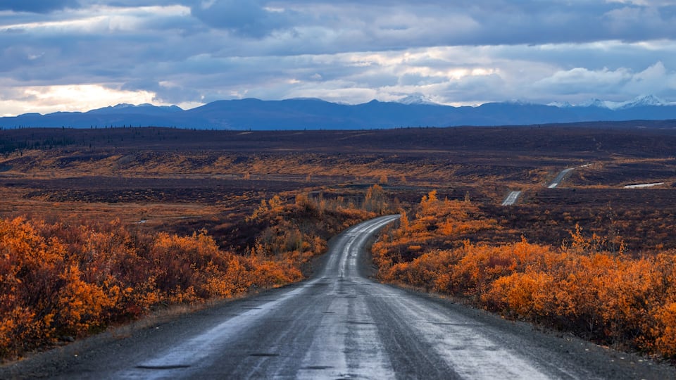 A winding road through a barren landscape with autumn-colored bushes and distant snow-capped mountains under a cloudy sky.