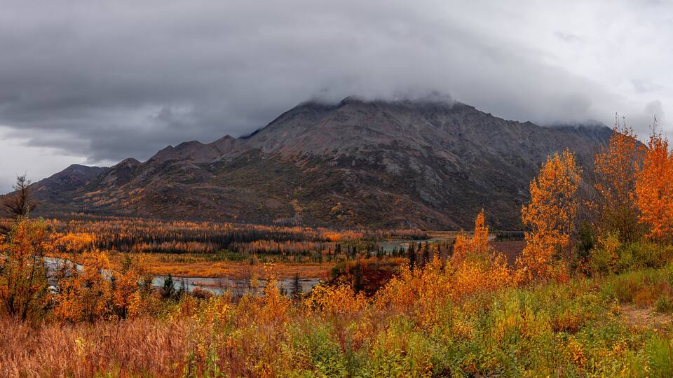 A mountainous landscape with autumn foliage, a river, and a cloudy sky.