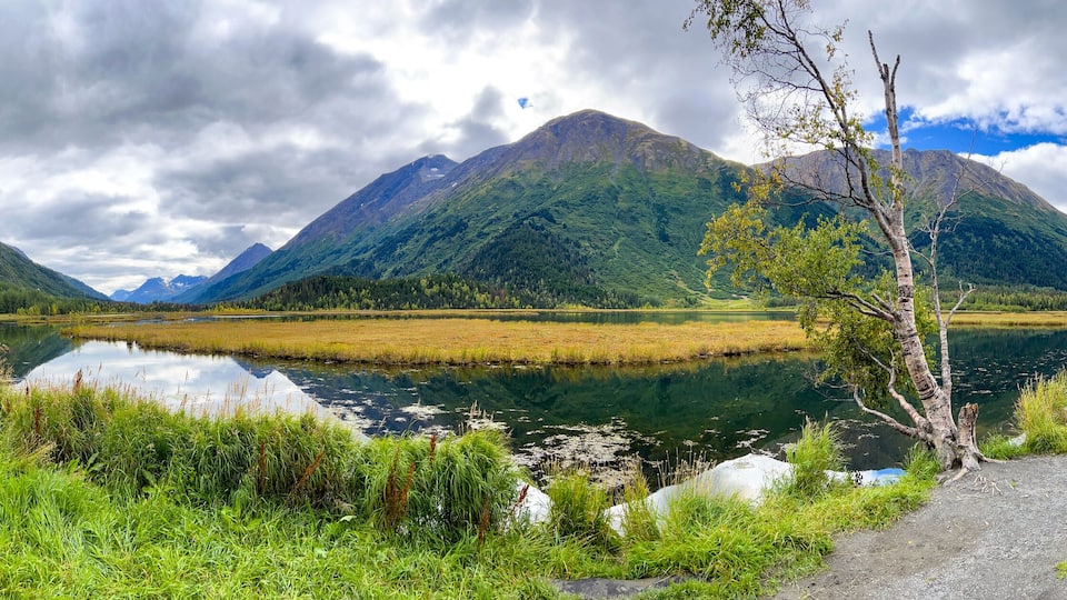 A mountain lake with a gravel path, surrounded by lush greenery and mountains.