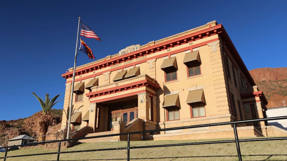 A historic courthouse with a red and beige facade, a flagpole with the American flag, and a stone wall in the foreground.