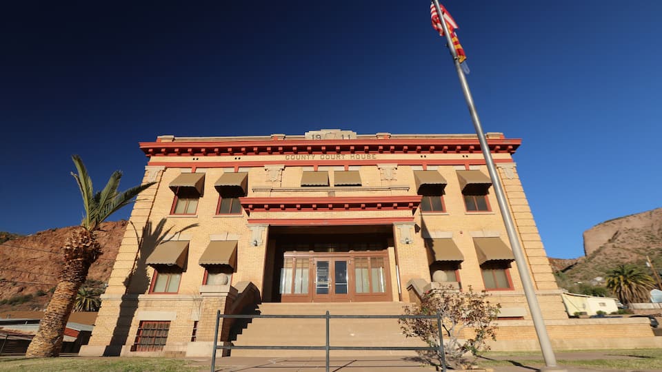 A courthouse with a red and beige facade, a flagpole with an American flag, and a palm tree in the foreground.