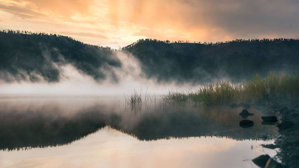 A calm lake with mist rising from the water, surrounded by forested hills and a sunset sky.