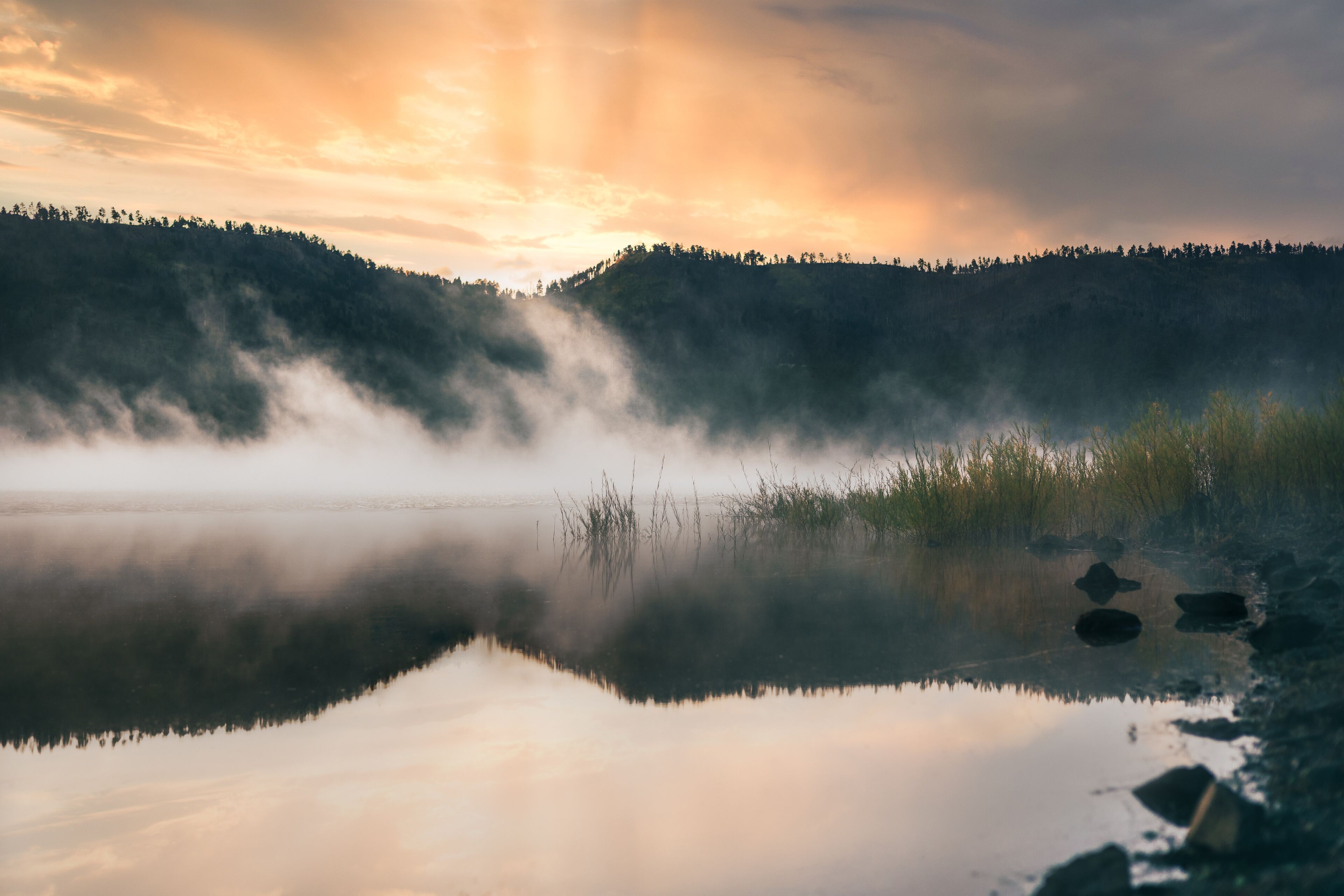 A calm lake with mist rising from the water, surrounded by forested hills and a sunset sky.