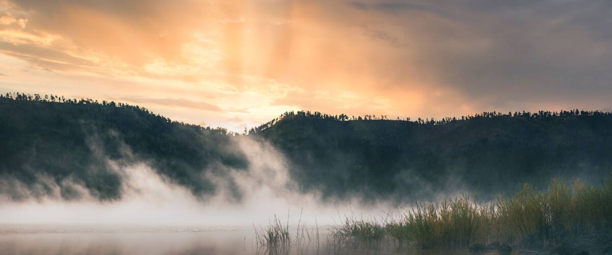 A calm lake with mist rising from the water, surrounded by forested hills and a sunset sky.