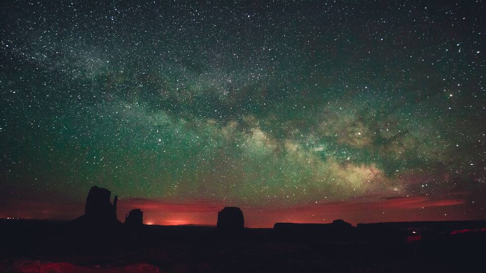 A starry night sky with the Milky Way visible, silhouetted rock formations, and a red glow in the distance.