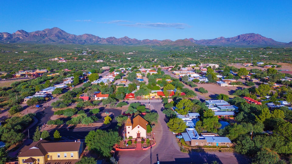 A small town with a central road, surrounded by houses and greenery, with mountains in the background.