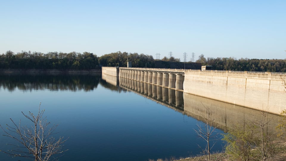 A dam with calm water reflecting its structure, surrounded by trees and a clear sky.