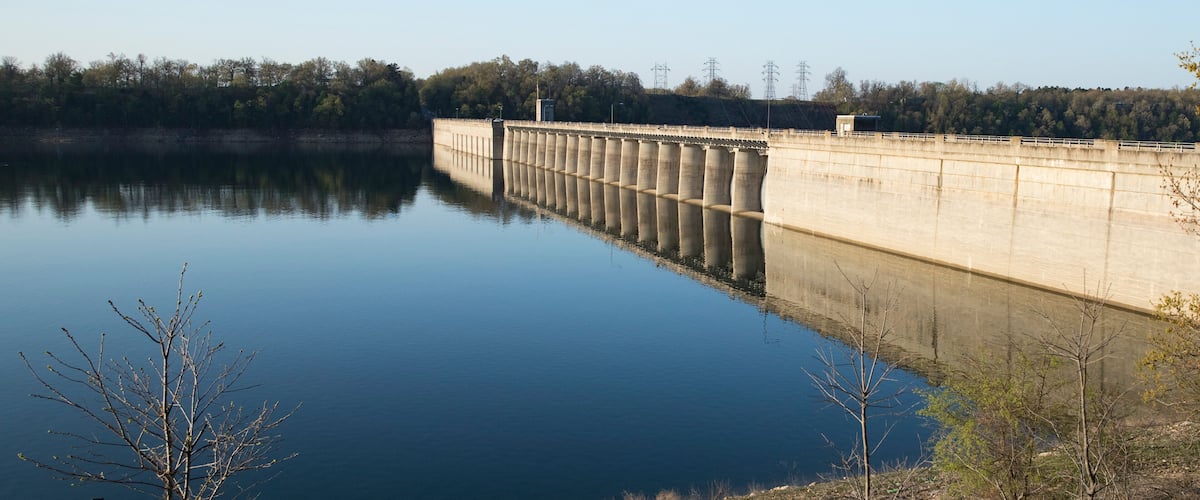 A dam with calm water reflecting its structure, surrounded by trees and a clear sky.
