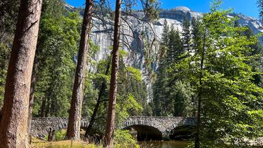 Un puente de piedra sobre un río en un bosque con árboles altos y una montaña rocosa al fondo.