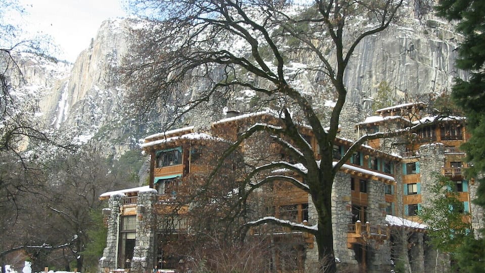 A snow-covered mountain lodge with stone walls and wooden balconies, surrounded by trees and a rocky cliff.